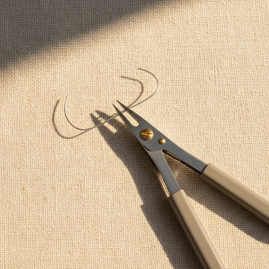 Close-up overhead shot of a reverse-action self-locking tweezer holding a thin silver wire in its tip, the wire visibly bowed slightly under the tweezer's spring tension. Workshop linen surface, warm directional light.